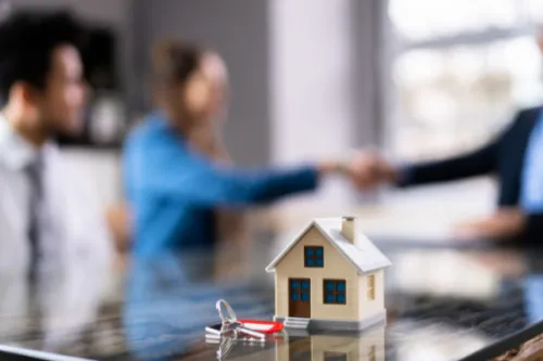 Intermediário de crédito a apoiar um casal na compra de casa, com chave e maquete de habitação sobre a mesa, representando o crédito habitação.